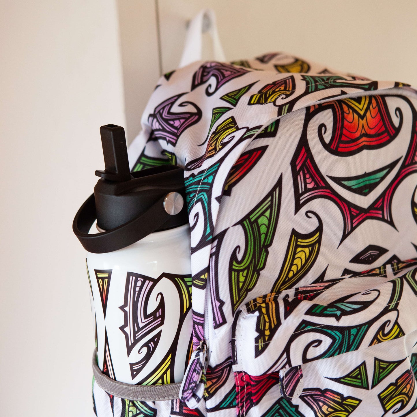 Close up of a Kids backpack showing the drink bottle in a pocket. The bag in white with colourful Maori design by Miriama Grace Smith.