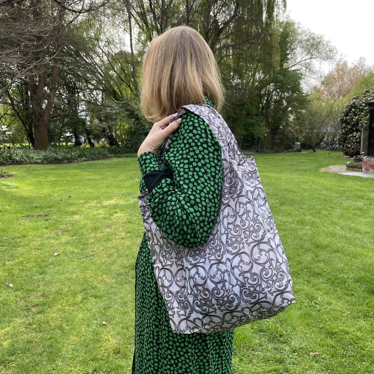 Woman standing in a garden in a black and green dress with her back to the camera. She has a black and white patterned reusable shopping tote bag over her shoulder.