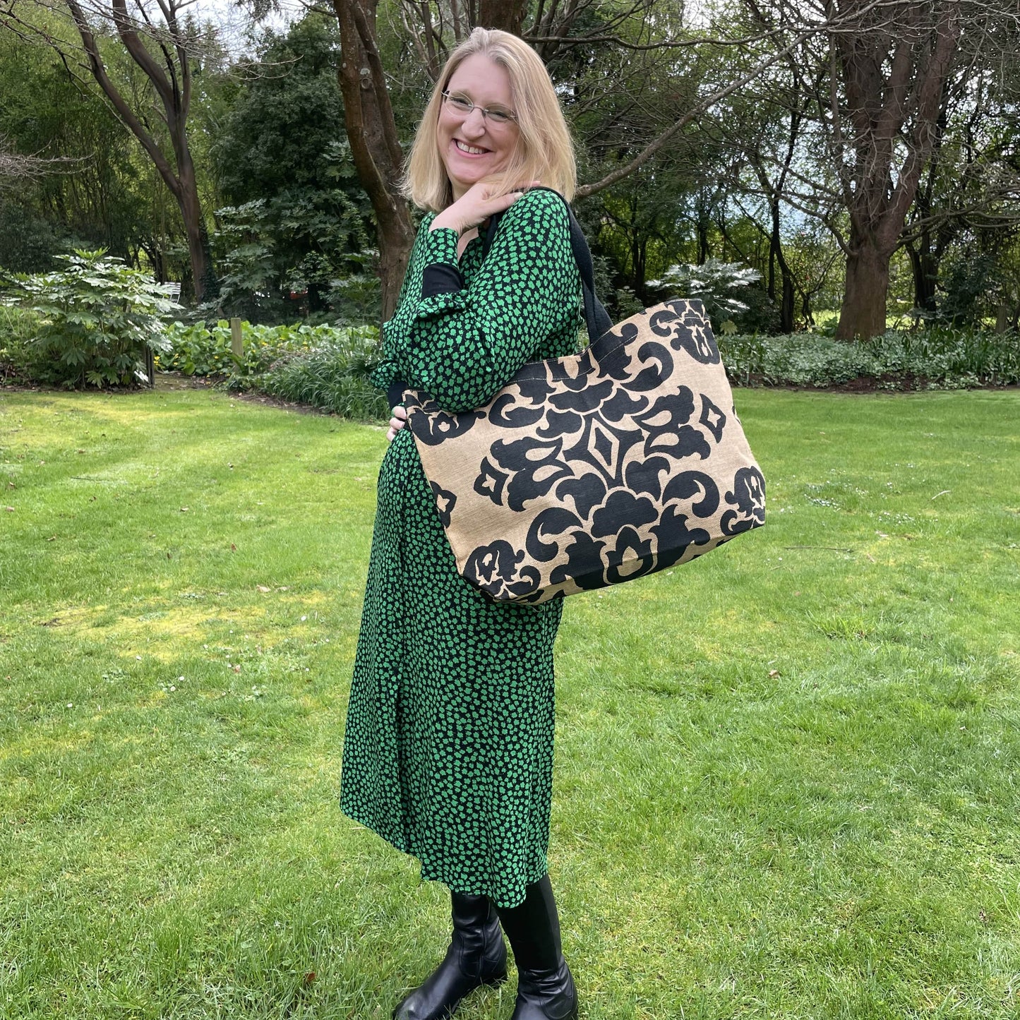 Blonde woman in a green and black dress holding a brown jute bag with black damask style print over her shoulder, in a lush green garden setting.