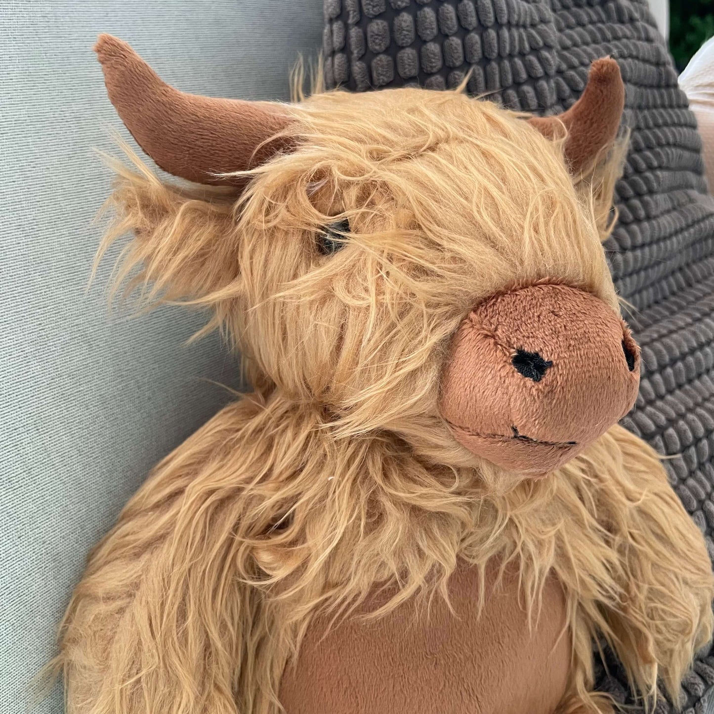 Close up of a soft toy Highland Cows face. It has a shaggy light brown fur, darker brown horns, nose and tummy.