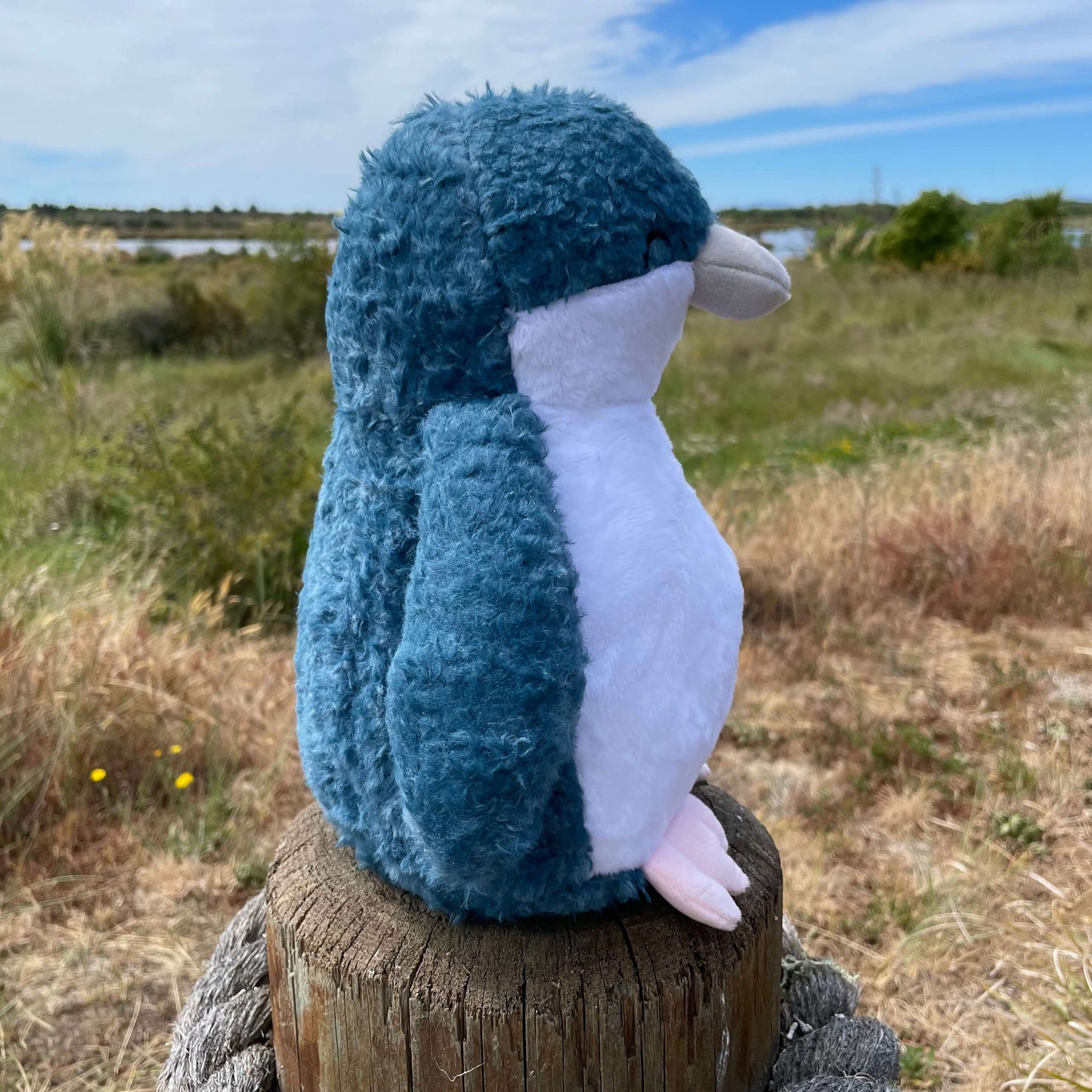Side view of a soft toy little blue penguin with white tummy and blue head and flippers sitting on a fence post with an estaury view in the background.