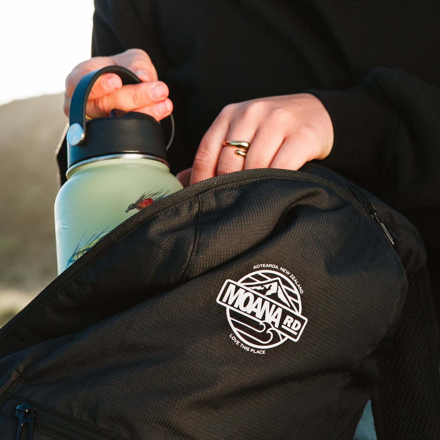 Close up of a person putting a drink bottle into a black backpack with white Moana RD logo.