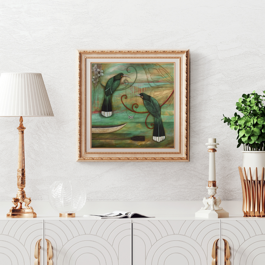 Art print by Kathryn Furniss of 2 green Huia birds with white tip tails sitting on linked Manuka branches styled like koru. Background features a green gold hue with a butterfly wing, waka and paddle. Framed in an ornate gold square frame, hanging on a white wall above an art deco style white credenza with gold fittings.