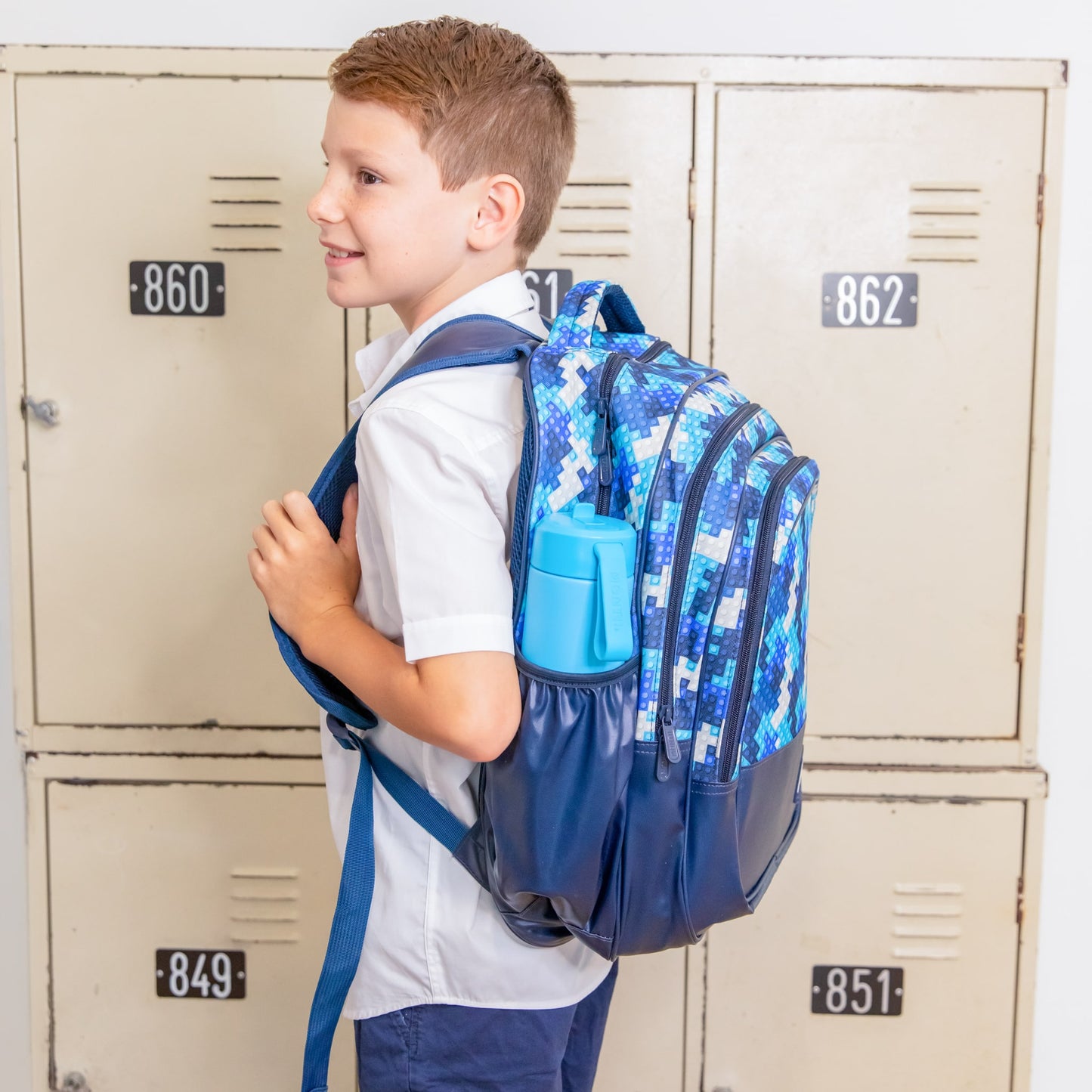 Young boy standing in front of a set of lockers with a Childrens backpack by brand Montiico that is multi shades of blue with a block print on the top half and navy blue on the bottom.