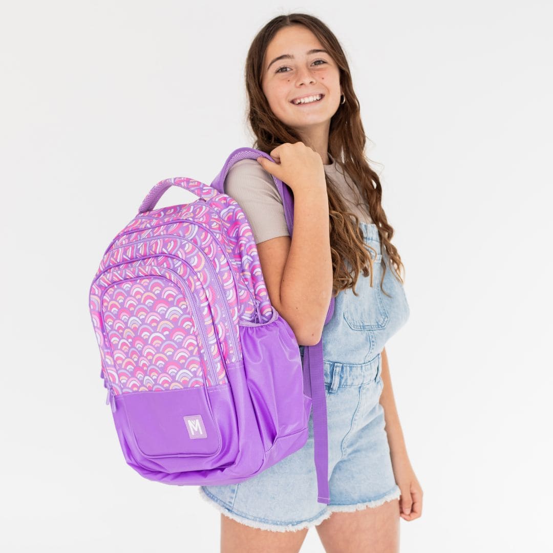 Young girl looking at the camera with a backpack over her shoulder. The backpack is purple with a peach, pink and white rainbow print.