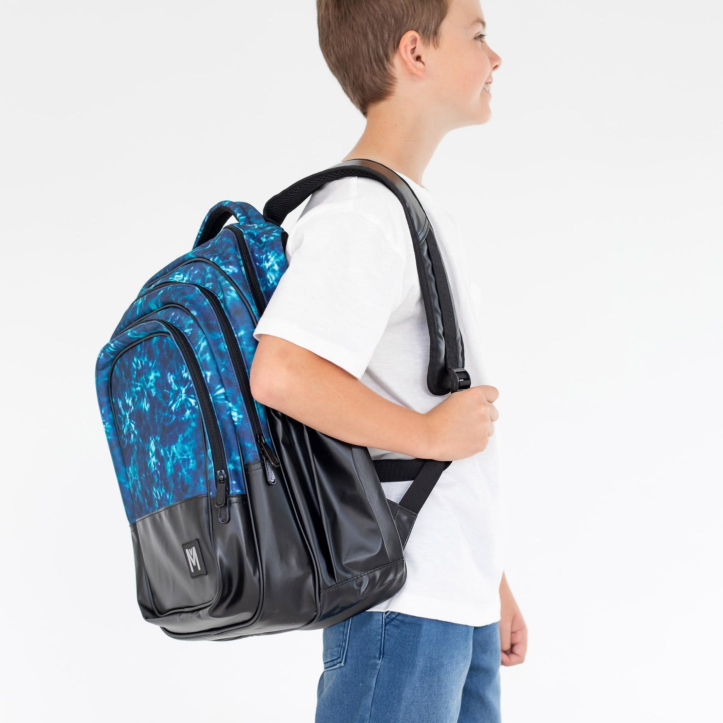 Young boy standing side on with a blue tie dye and black print backpack over his shoulder.