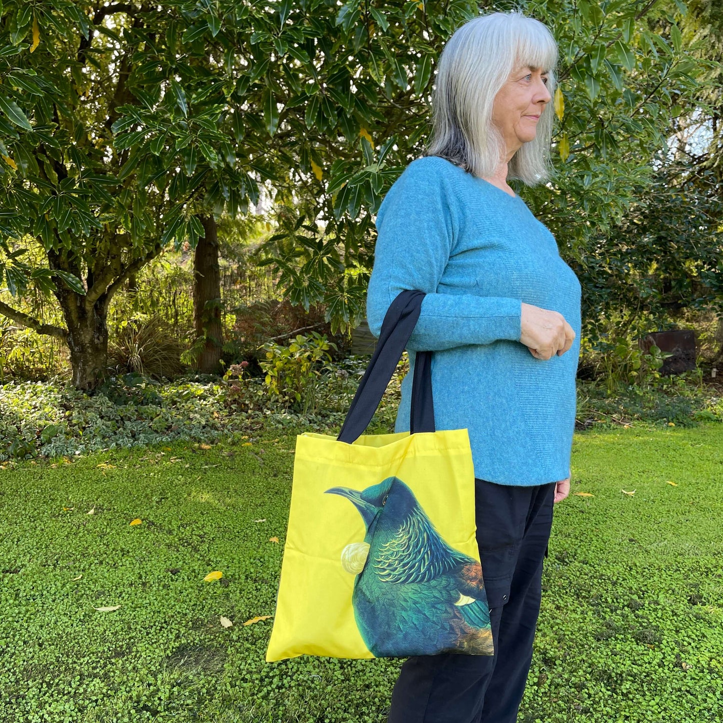 Woman with a tote bag over her arm. The bright yellow tote features a Tui bird.