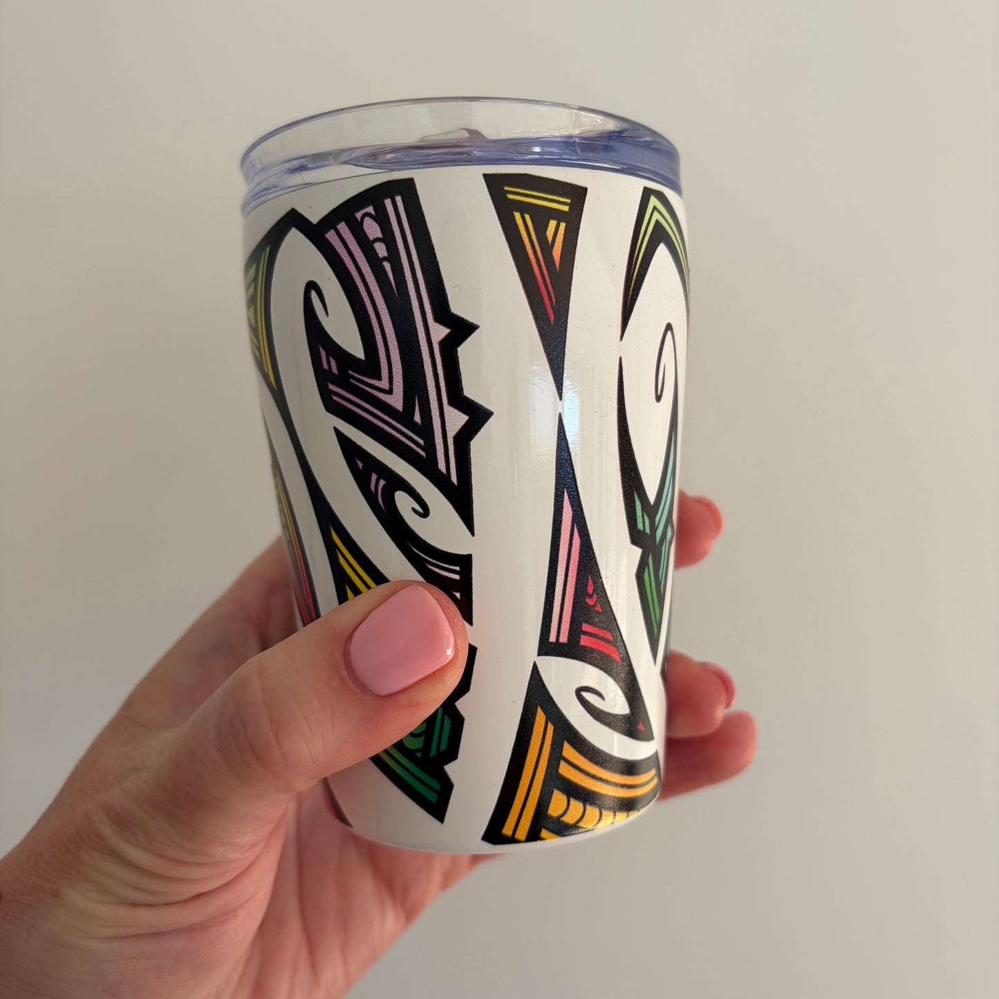 A hand with pink nail polish holds a white insulated tumbler featuring a colourful Māori tribal pattern in black, green, yellow, and pink, set against a plain light gray background.
