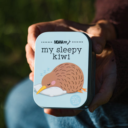 A person holds a small square tin labeled 'MOANA RD  my sleepy kiwi' against an outdoor blurred background. The tin has a light blue background with a cartoon kiwi sleeping on a white pillow.