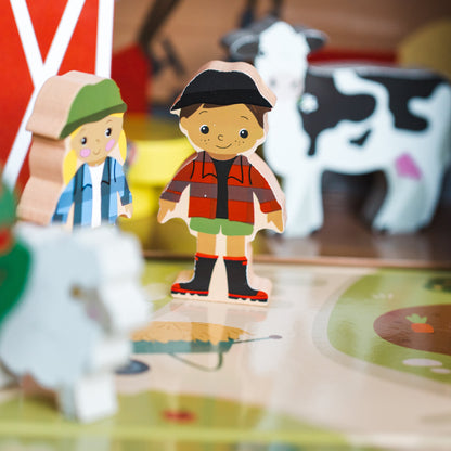 Wooden farm-themed toy set with small cutout character figures and animal pieces arranged on a play mat, with a blurred barn and cow in the background.