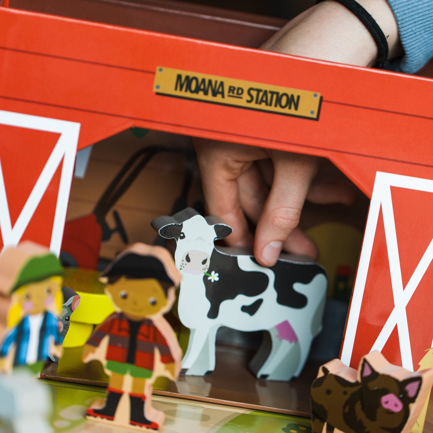 A close-up of a person's hands playing with a wooden toy barn set labeled 'MOANA RD STATION'. The red and white barn contains wooden farm animal and character figures, including a black-and-white cow, a pig, and small cutout people, set on a wooden floor.