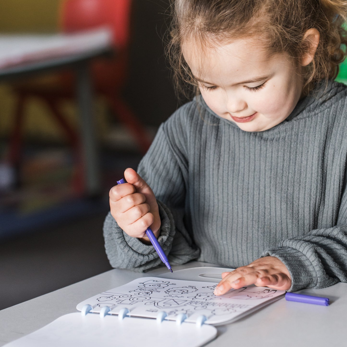 A young girl with her hair in a ponytail is sitting at a table, coloring in a book with a purple pen. She is wearing a gray ribbed sweater, and the background shows a softly blurred indoor setting.