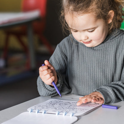 A young girl with her hair in a ponytail is sitting at a table, coloring in a book with a purple pen. She is wearing a gray ribbed sweater, and the background shows a softly blurred indoor setting.