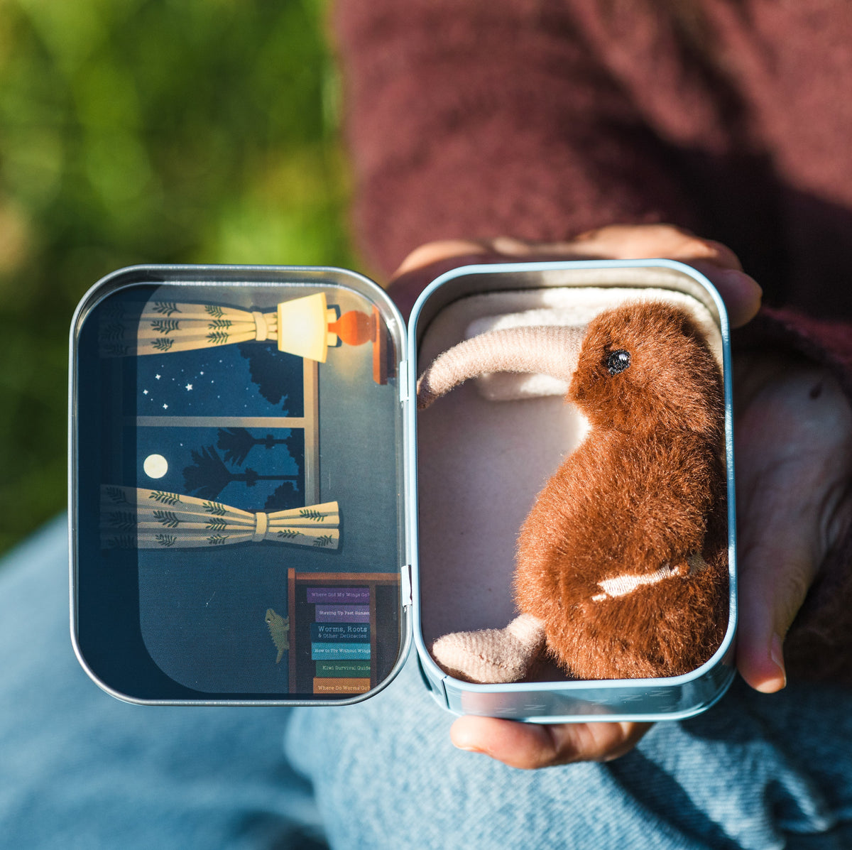A person outdoors holds an open square tin containing a small brown plush kiwi bird. The left side of the tin shows a miniature illustrated night scene with a window, stars, and curtains.
