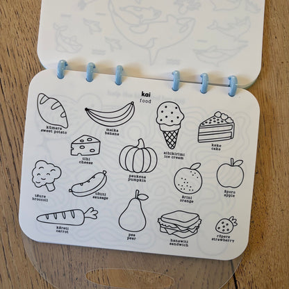A children's activity colouring book page featuring black line drawings of fruits, vegetables, and snacks on a white page, held open by light blue binder rings on a wooden surface.