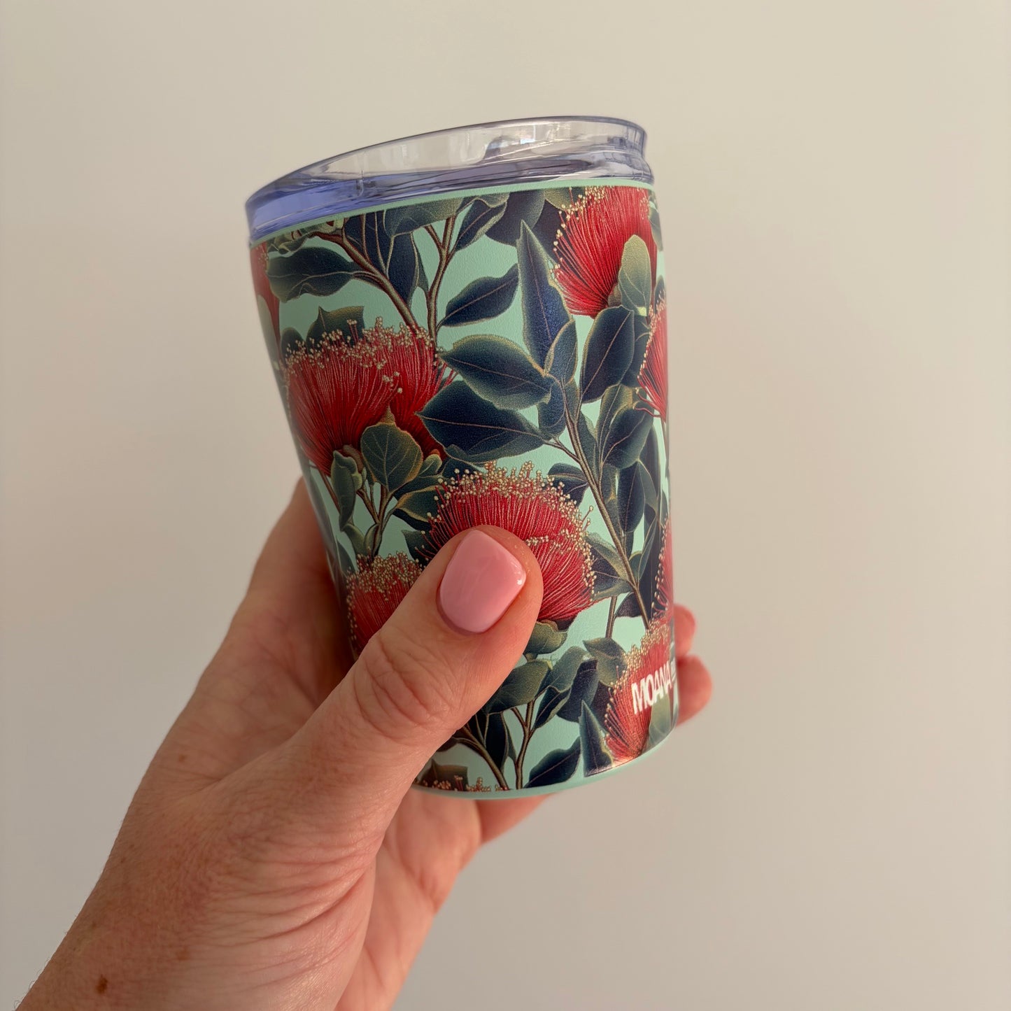 A hand holding a clear-lidded stainless tumbler with a botanical print featuring red pohutukawa flowers and dark green leaves on a pale mint background.