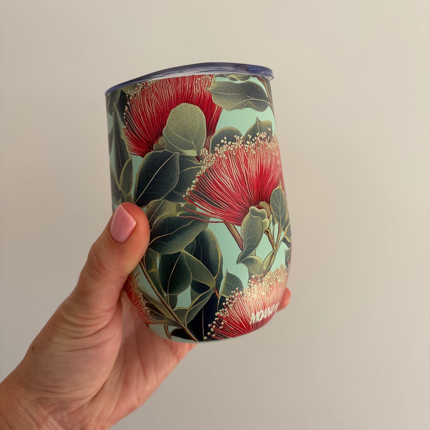 A hand holding a reusable tumbler with a floral print featuring red pōhutukawa flowers and green leaves on a pale mint background, set against a plain light gray wall.