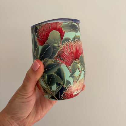 A hand holding a reusable tumbler with a floral print featuring red pōhutukawa flowers and green leaves on a pale mint background, set against a plain light gray wall.