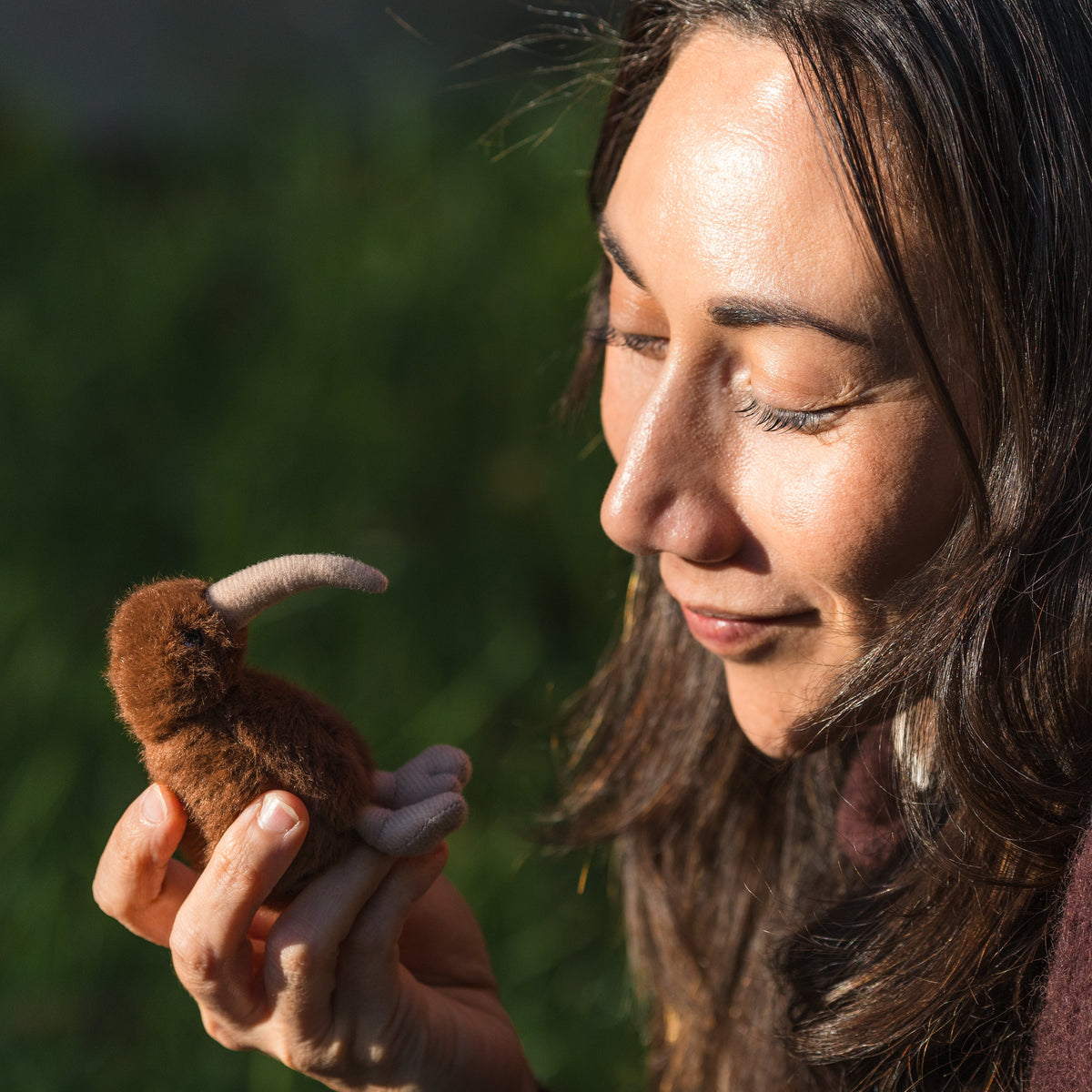 A woman outdoors holds a small brown plush Kiwi bird toy with a curved cream-colored beak in her hands, with a blurred green natural background.