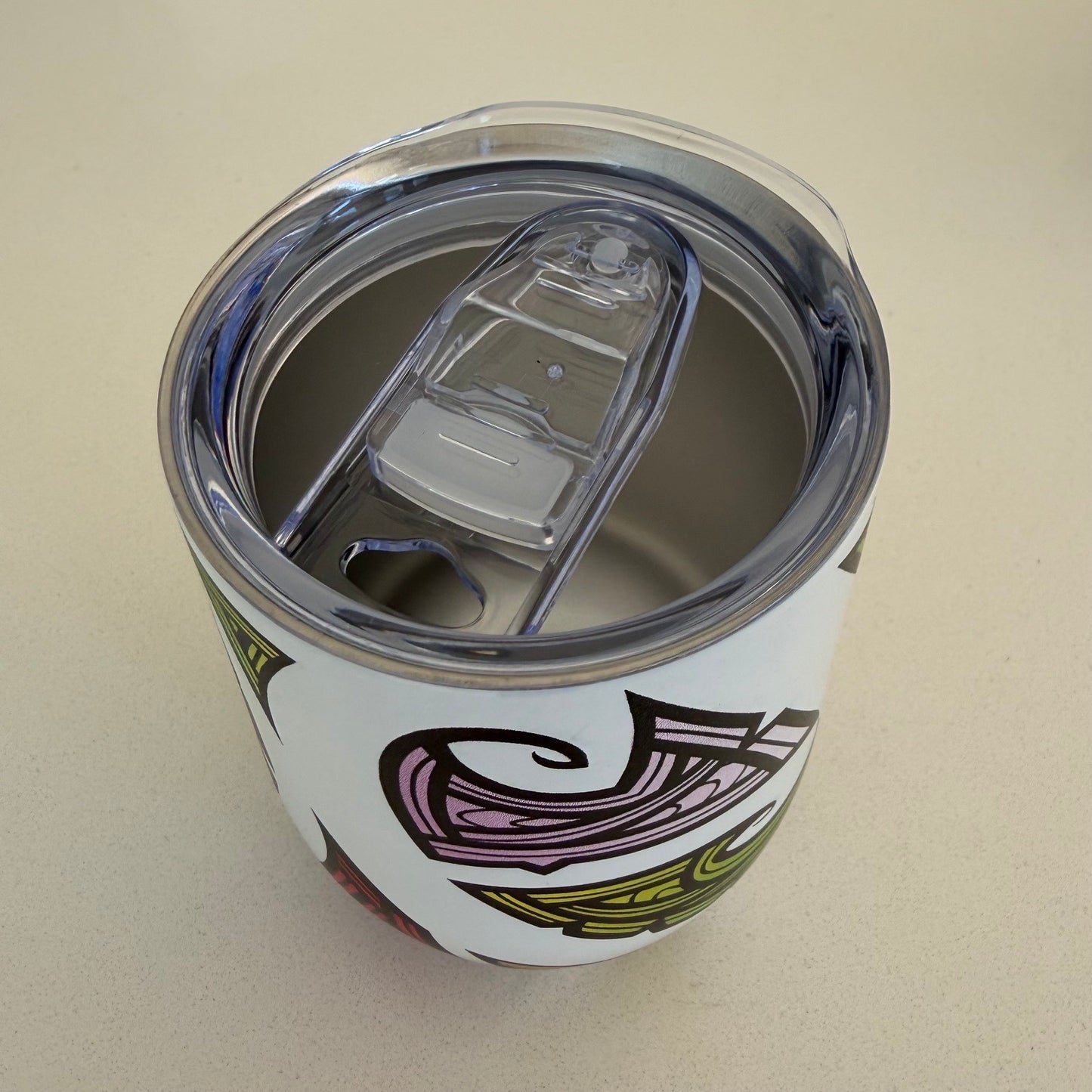 A white insulated tumbler with a clear lid and a colorful tribal-style graphic, shown from a top-down angle on a light beige surface.