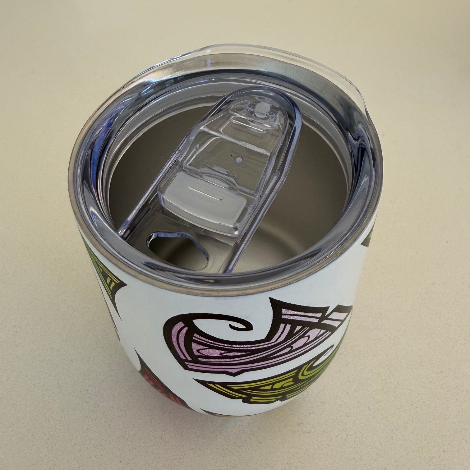 A white insulated tumbler with a clear lid and a colorful tribal-style graphic, shown from a top-down angle on a light beige surface.