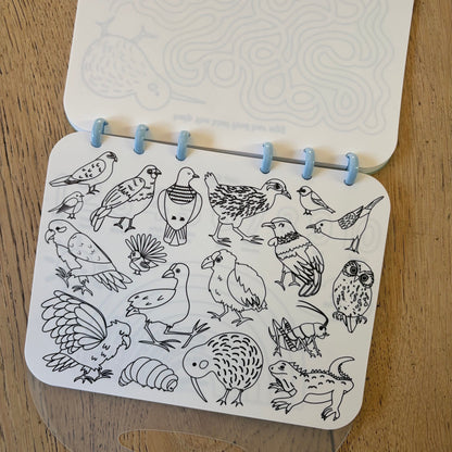 A white colouring book with rounded corners, bound by light blue ring loops, lying open on a wooden surface. The shown page is filled with black line drawings of various New Zealand birds.