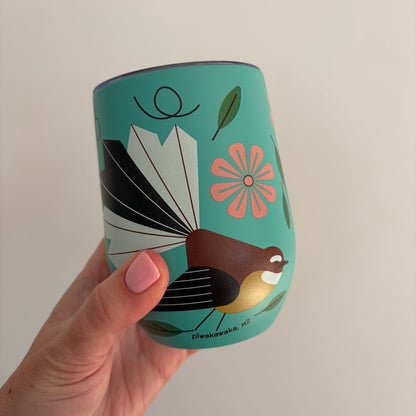 A hand with pink nail polish holds a teal insulated tumbler featuring a stylized Fantail bird and floral illustration. The tumbler has a clear lid and is photographed against a plain light background.