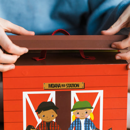 A child's hands open a red gift box designed like a barn, labeled 'MOANA RED STATION,' with illustrated children on the front. The child is wearing a blue shirt and sitting indoors.