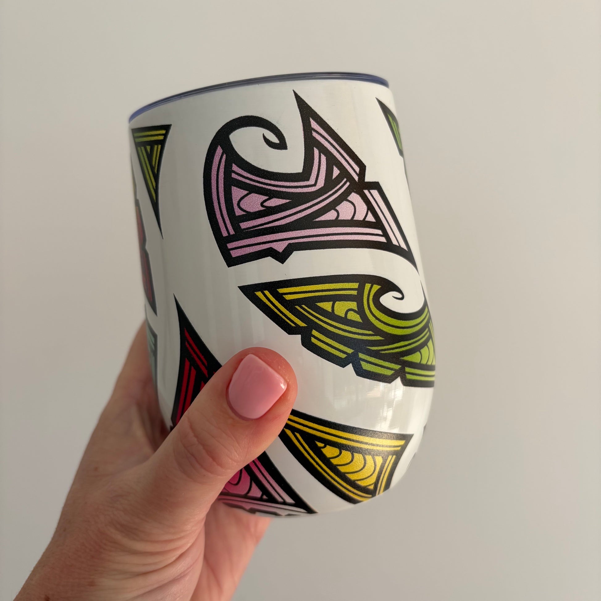 A hand with pink nail polish holds a white stainless tumbler decorated with colourful geometric Maori tribal-style patterns in green, pink, and yellow.