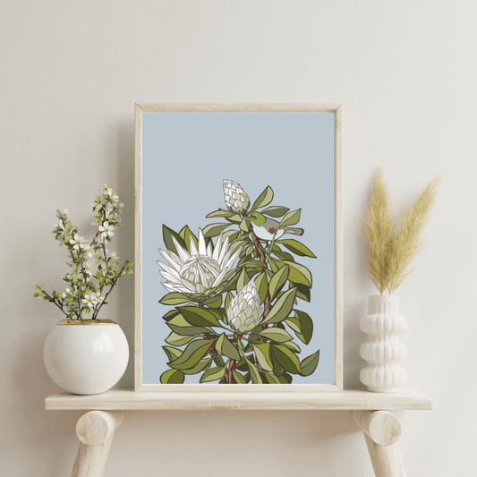 Digital print art by Bridget King featuring a Tahou bird amongst white Protea flowers. Framed in a light wooden frame sitting on a wooden side table with a white vase either side containing flowers.