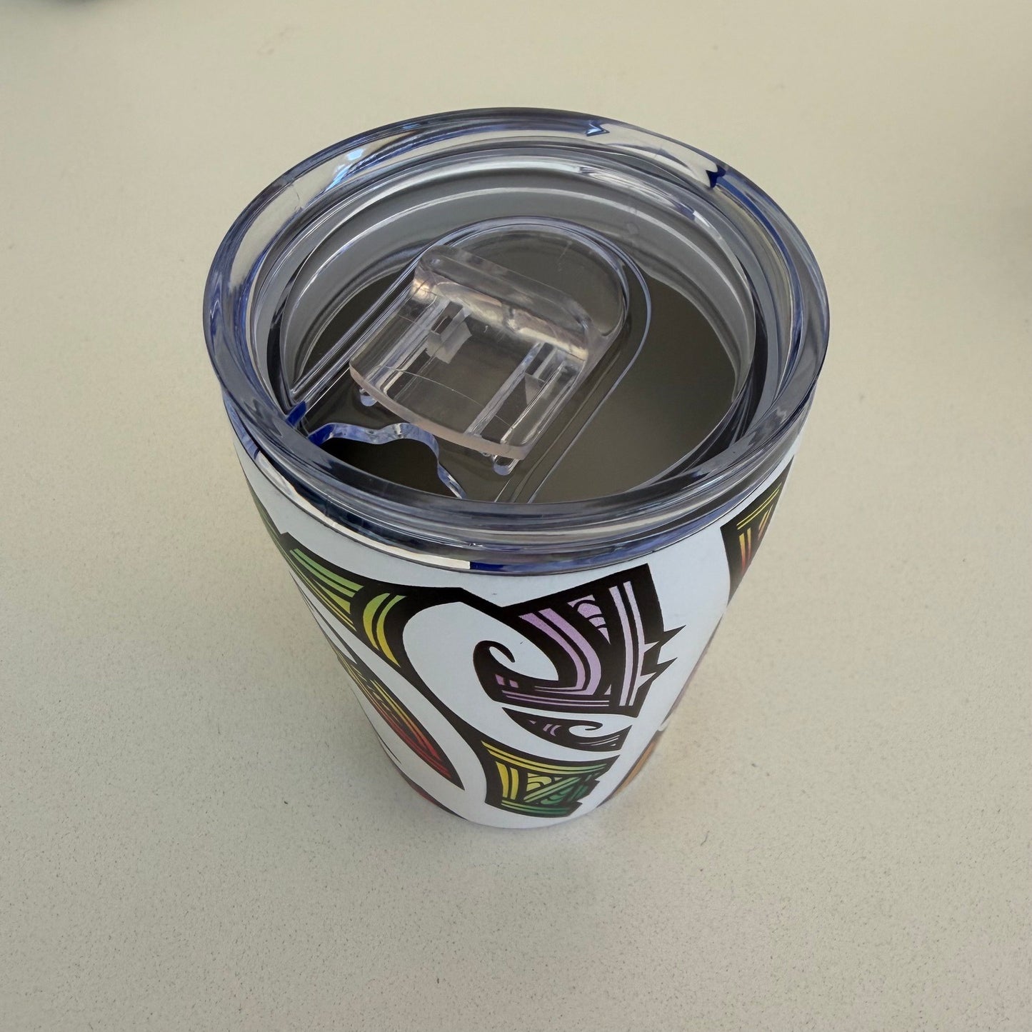 A clear plastic lid on a stainless insulated tumbler and a white body featuring a bold black, green, and purple tribal-style graphic, shown on a light beige surface.