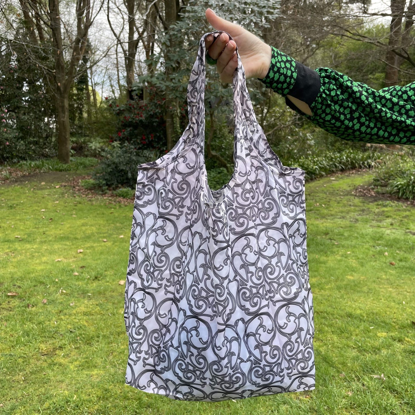 Womans hand holding a patterned black and white reusable shopping tote bag with a lush green garden in the background.