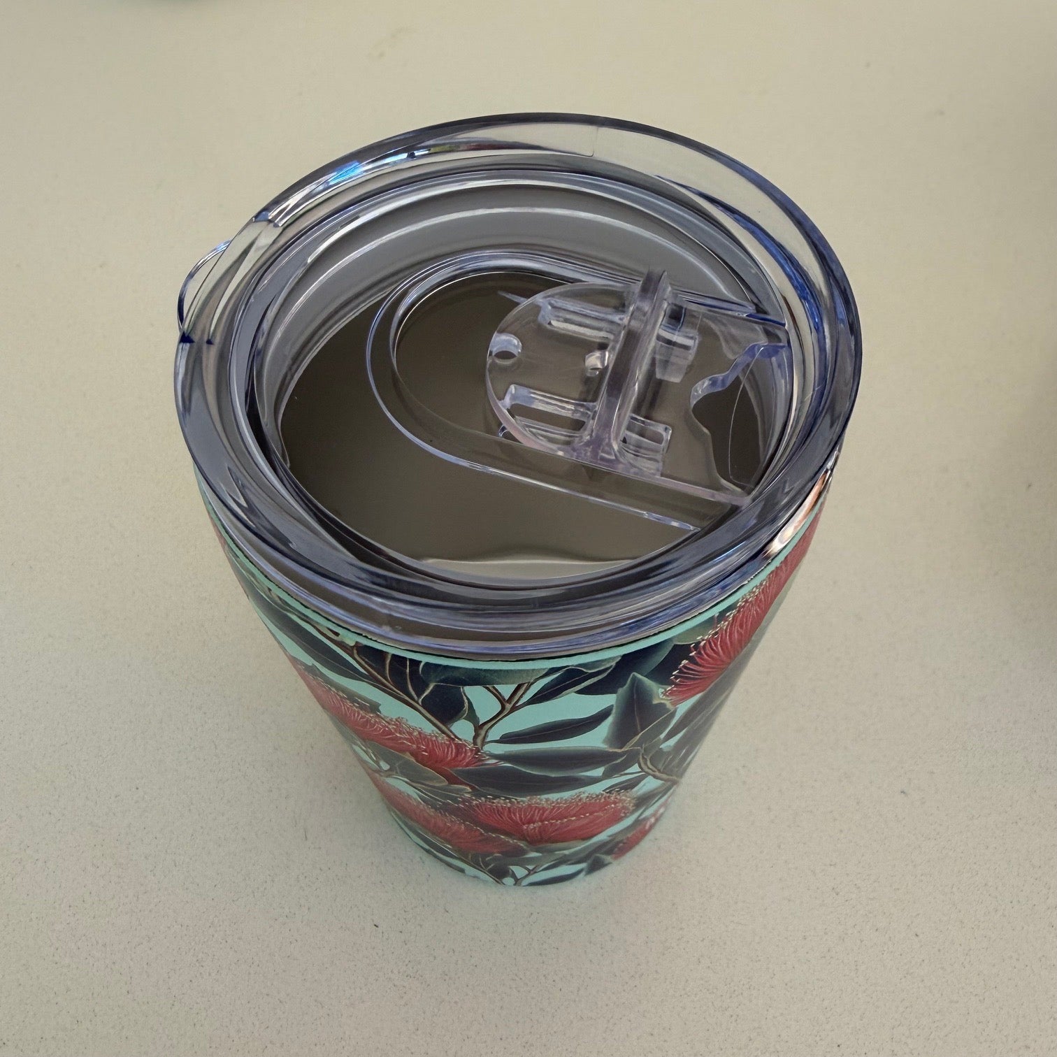 A clear plastic lid on a stainless travel tumbler, featuring a printed red pohutukawa floral pattern with green leaves on a light background.