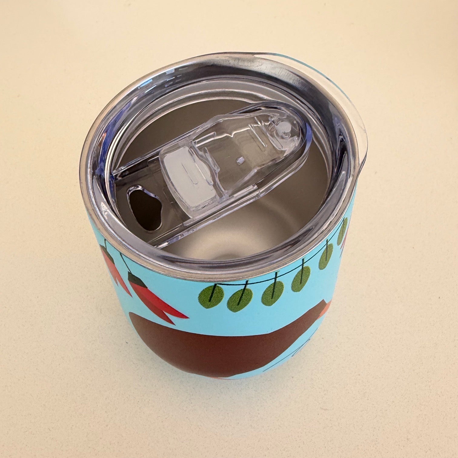 A close-up of a stainless steel travel tumbler with a clear lid and flip-up straw, decorated with a light blue background and brown kiwi bird design featuring red kaka flowers and green leaves.