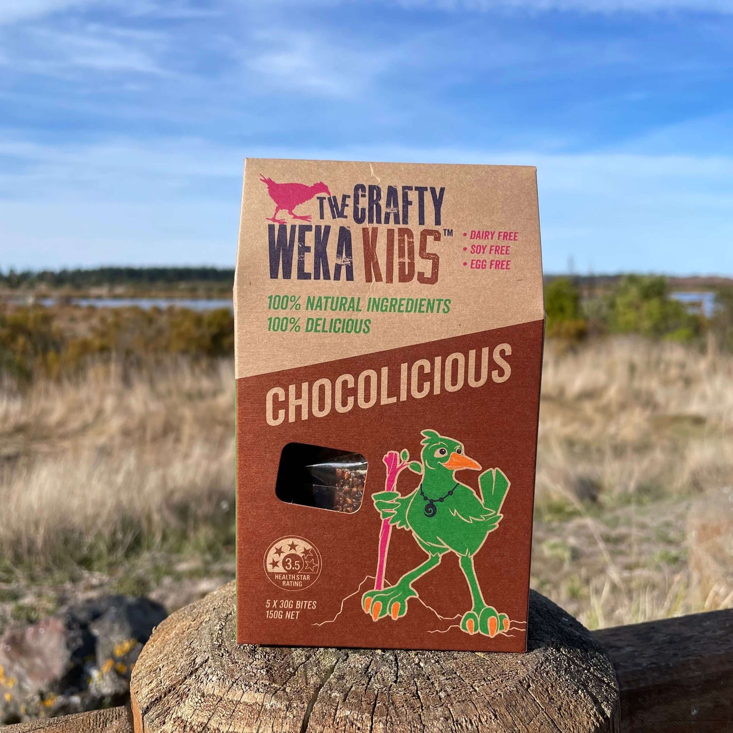 Cardboard pouch of chocolicious muesli bars from Crafty Weka sitting on a fence post overlooking an Estuary.