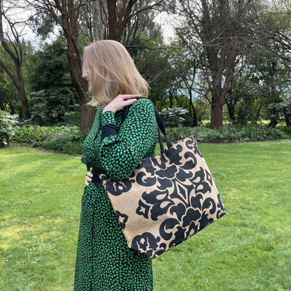 Close up of a jute tote bag with black damask style print over the shoulder of a woman in a black and green dress amongst a green garden setting.