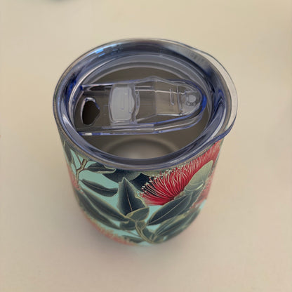 A clear plastic flip top lid on a stainless travel tumbler, featuring a red pohutukawa floral print with green leaves shown on a light beige background.