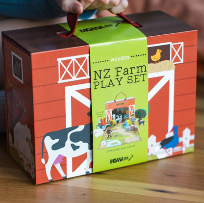 A person's hand holding a red handle of the 'Māwha wooden NZ Farm Play Set' box, which is decorated with illustrations of a barn, cow, and chicken, sitting on a wooden floor.