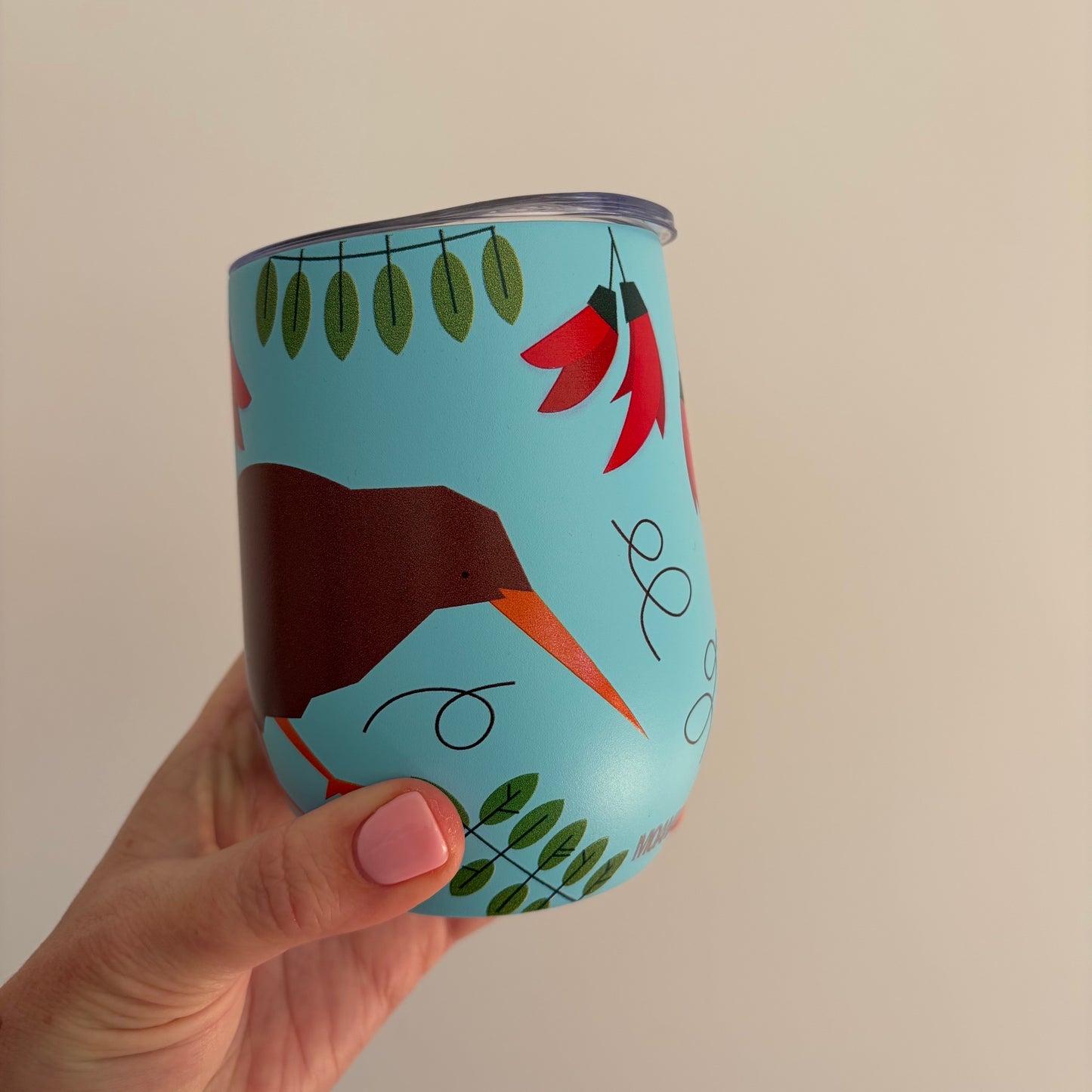 A hand with pink nail polish holds a light blue insulated tumbler featuring a stylized brown kiwi bird, red kaka flowers, green leaves, and black abstract line art against a plain beige background.
