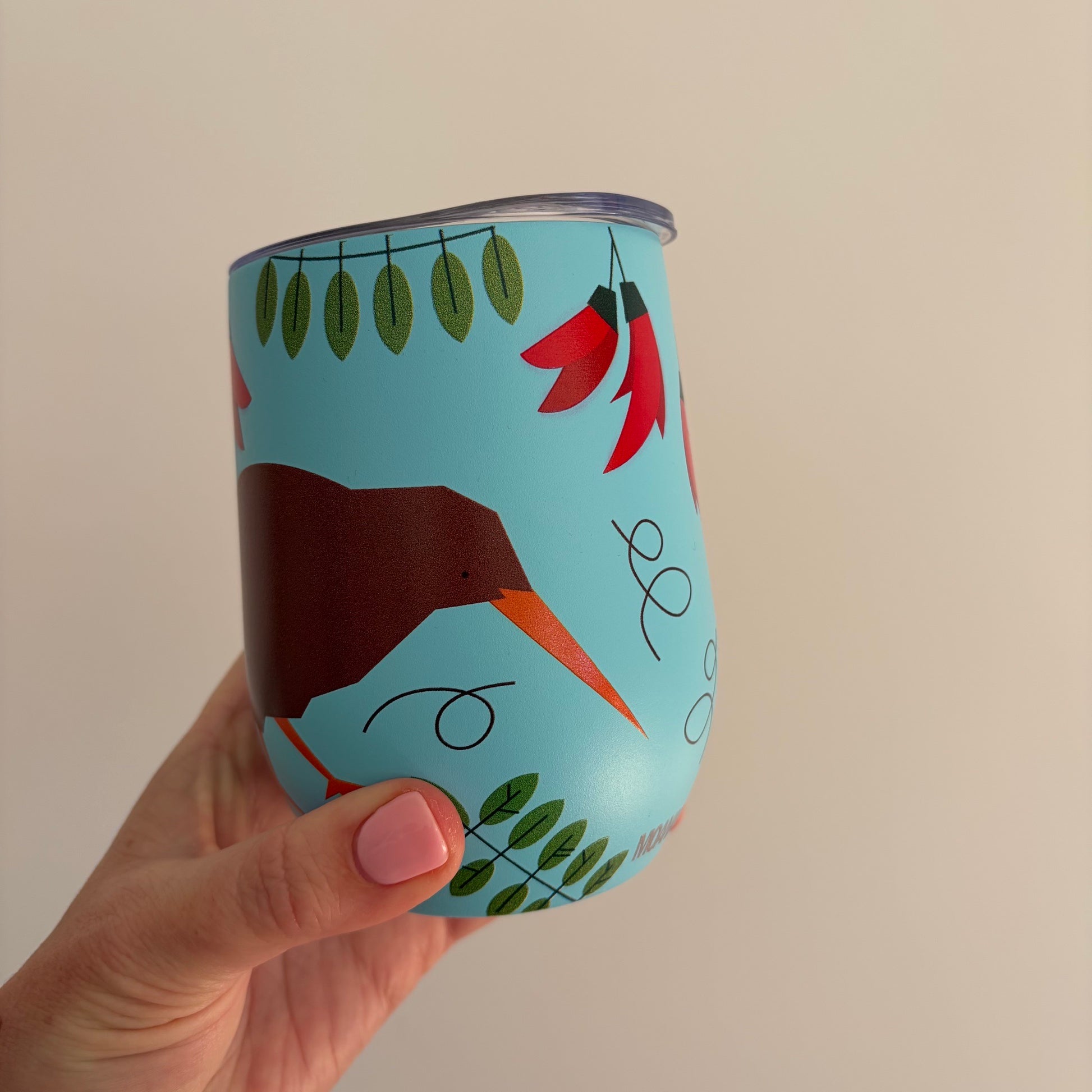 A hand with pink nail polish holds a light blue insulated tumbler featuring a stylized brown kiwi bird, red kaka flowers, green leaves, and black abstract line art against a plain beige background.