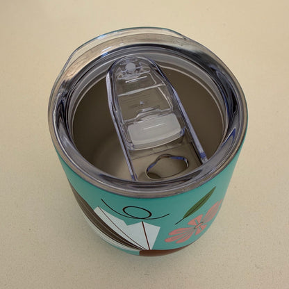A close-up top-down view of a teal insulated tumbler with a clear lid and flip spout, decorated with a pink flower, Fantail bird and green leaf design, on a light beige background.