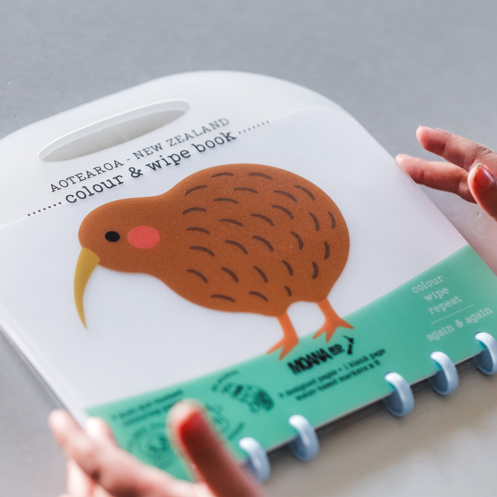 A children's spiral-bound book titled 'Aotearoa - New Zealand colour & wipe book' being held by two hands. The cover features a large kiwi bird illustration on a white background with a mint green bottom section.