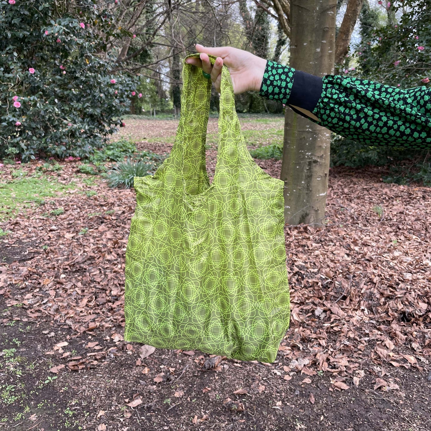 Womans hand holding a swirly green patterned reusable shopping tote bag with autumn leaves and trees in the background.