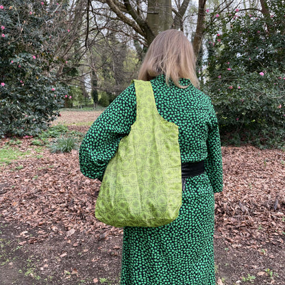 Woman standing in a bare autum garden in a black and green dress with her back to the camera. She has a swirly green patterned reusable shopping tote bag over her shoulder.