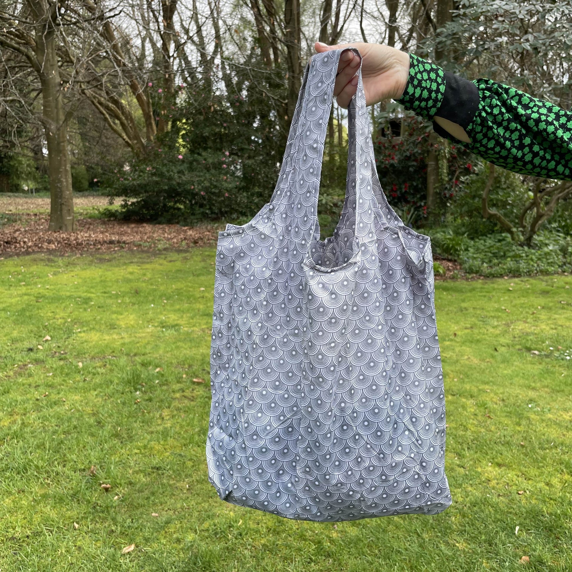 Womans hand holding a circle patterned grey and white reusable shopping tote bag with a lush green garden in the background.