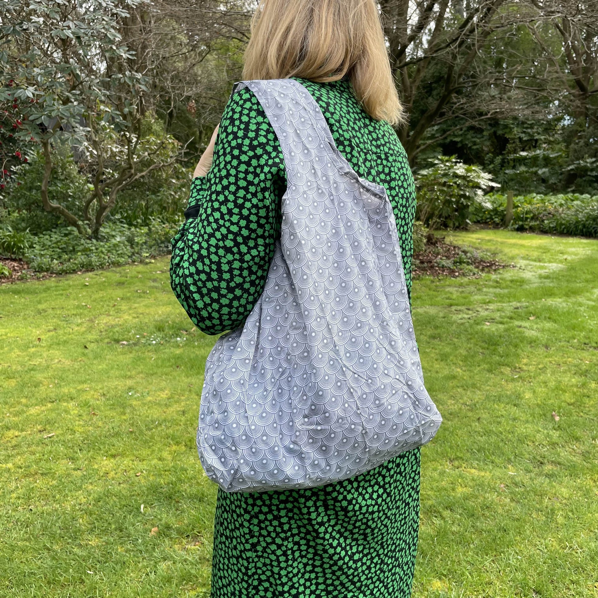 Woman standing in a garden in a black and green dress with her back to the camera. She has a grey and white circle patterned reusable shopping tote bag over her shoulder.