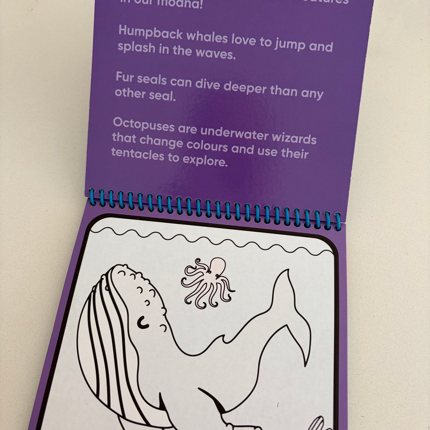 A children's spiral-bound booklet or activity book shown open on a light beige surface. The page has a purple header with printed text about whales, seals, and octopuses, and a white lower panel with black line drawings of a whale and octopus.