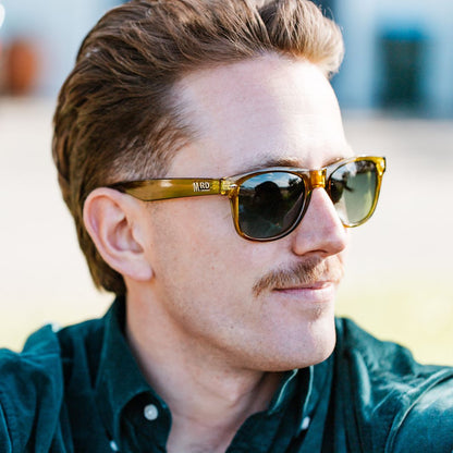 A man wearing olive green acetate sunglasses with dark lenses and a green button-down shirt, photographed in a bright outdoor setting with a blurred background.