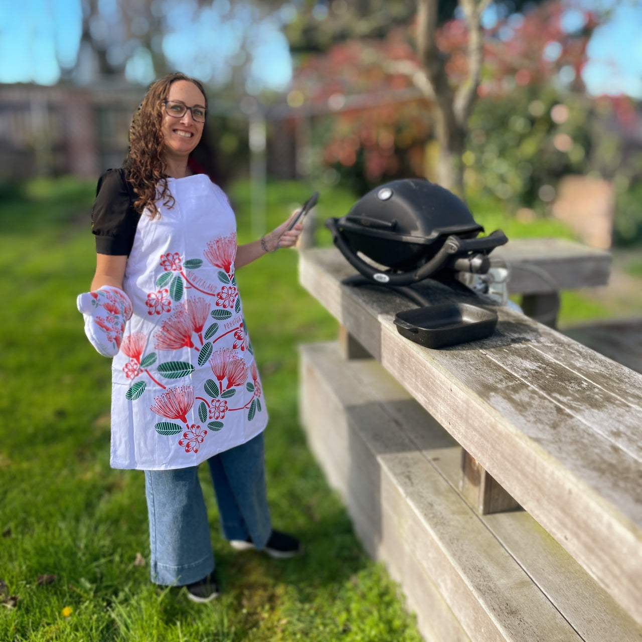 Woman wearing a white apron with red and green pohutukawa flower print at a BBQ.