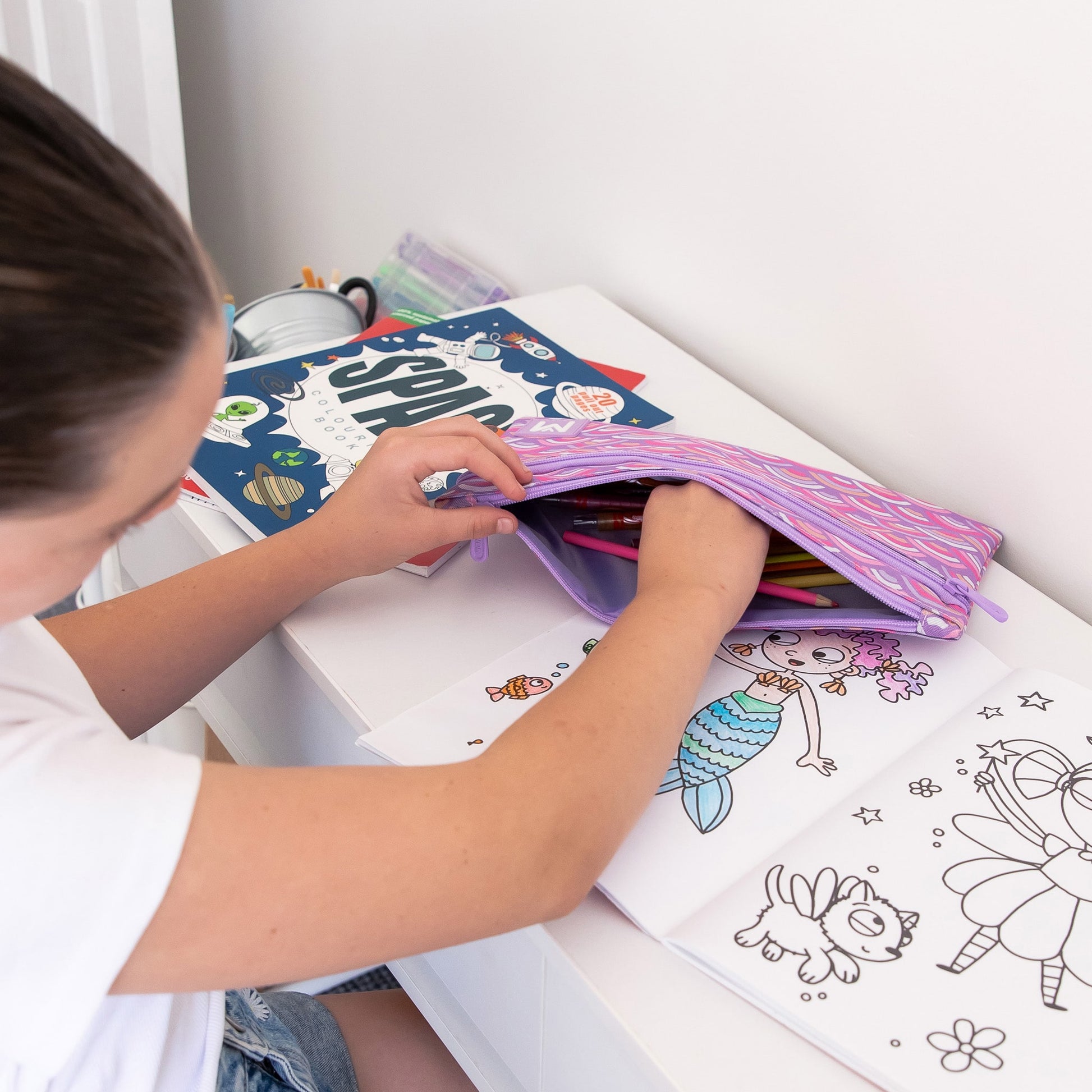 Child sitting at a desk with colouring pages and reaching into a purple pencil case full of colouring pencils.